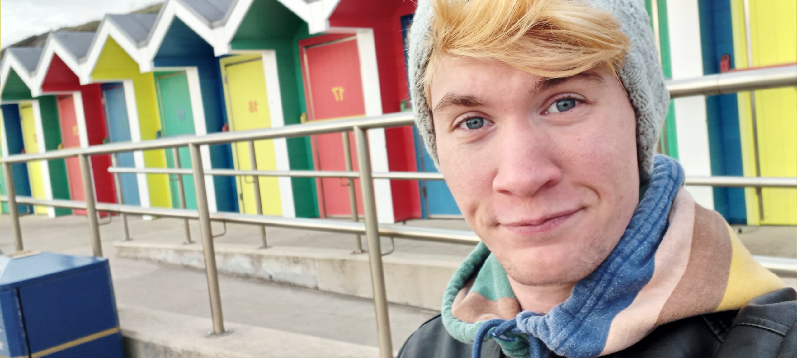 Riley, a white nonbinary person with short bleach blonde hair, smiles while standing in front of the Barry Island beach huts.